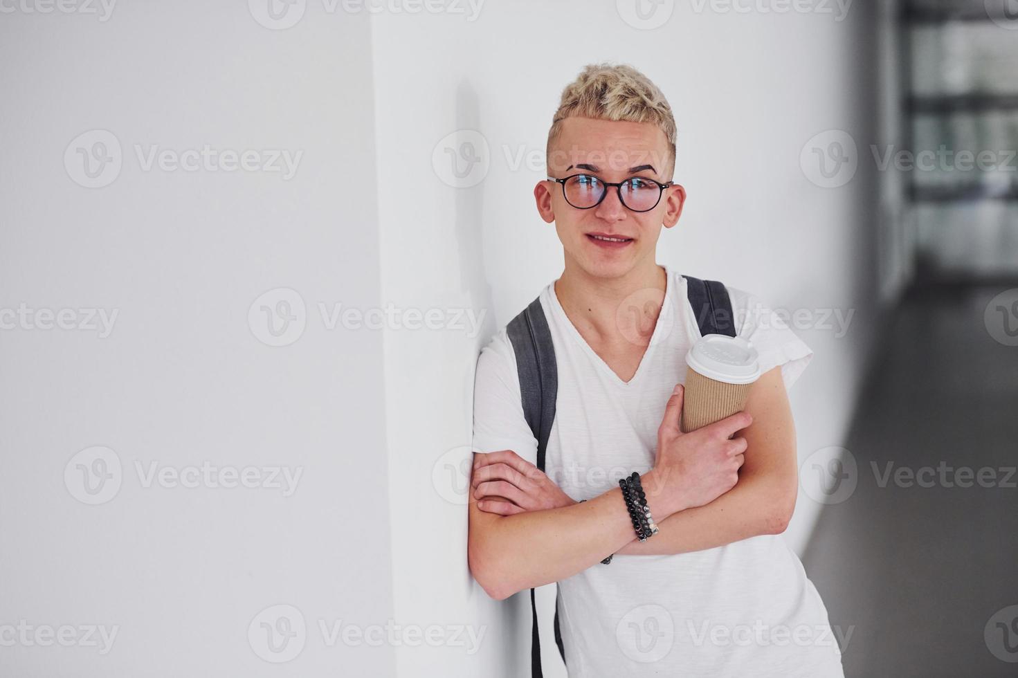 étudiant en vêtements décontractés et avec sac à dos se tient à l'intérieur contre un mur blanc avec une tasse de boisson photo