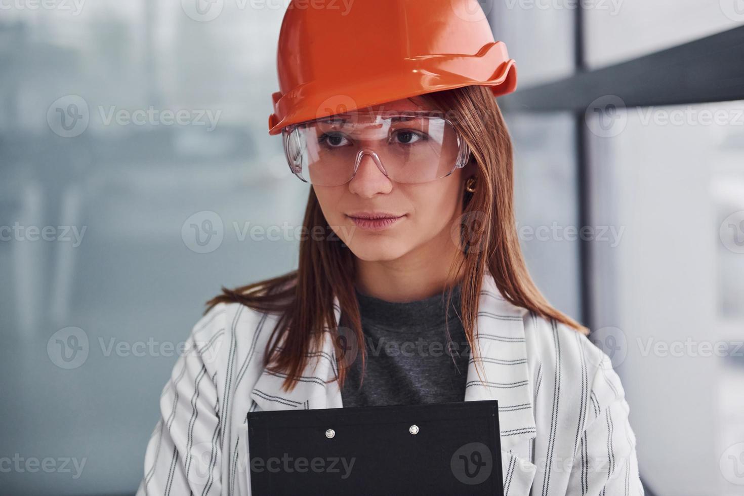 portrait de jeune travailleuse avec casque de protection, lunettes et bloc-notes. debout au bureau photo