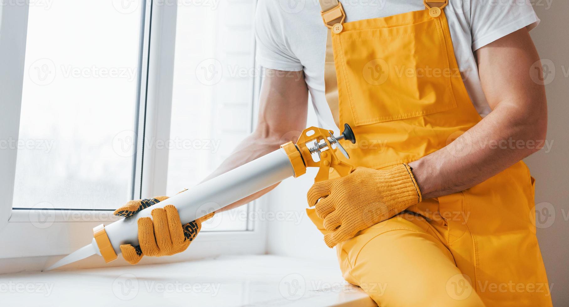 homme à tout faire en uniforme jaune travaille avec de la colle pour fenêtre à l'intérieur. conception de rénovation de maison photo