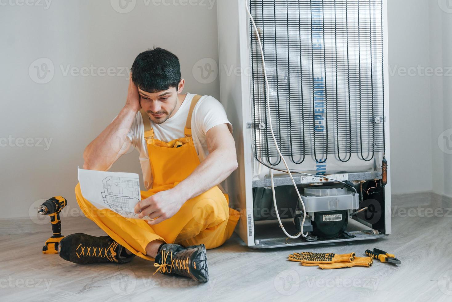 bricoleur confus en uniforme jaune manuel de lecture pour réfrigérateur à l'intérieur. conception de rénovation de maison photo