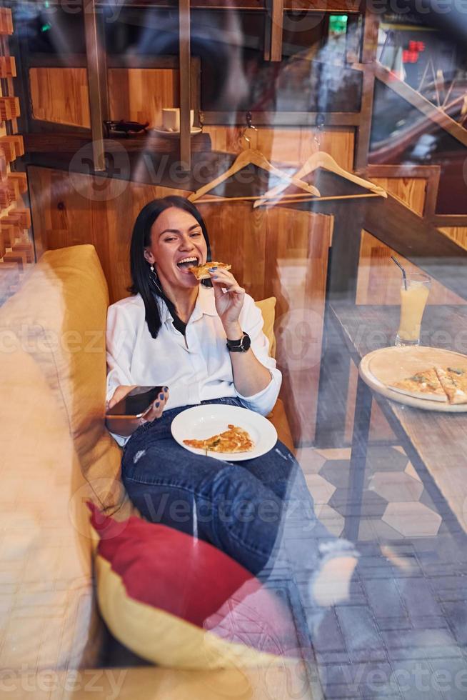 jeune brune assise à l'intérieur avec une pizza et un smartphone à la main photo