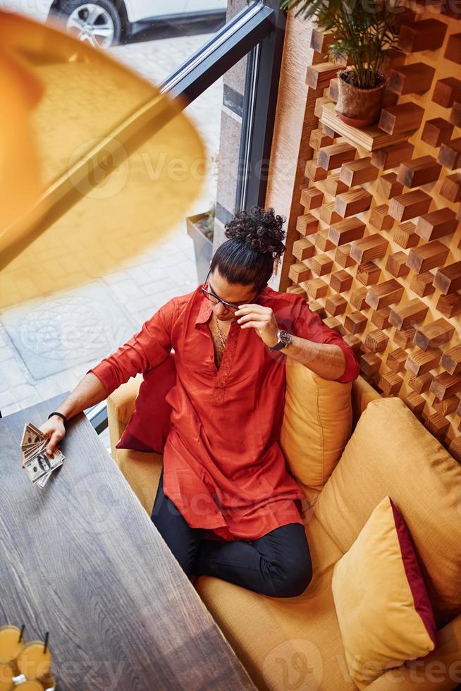 vue de dessus d'un homme riche en vêtements rouges et noirs assis à l'intérieur dans un café avec beaucoup d'argent en main photo