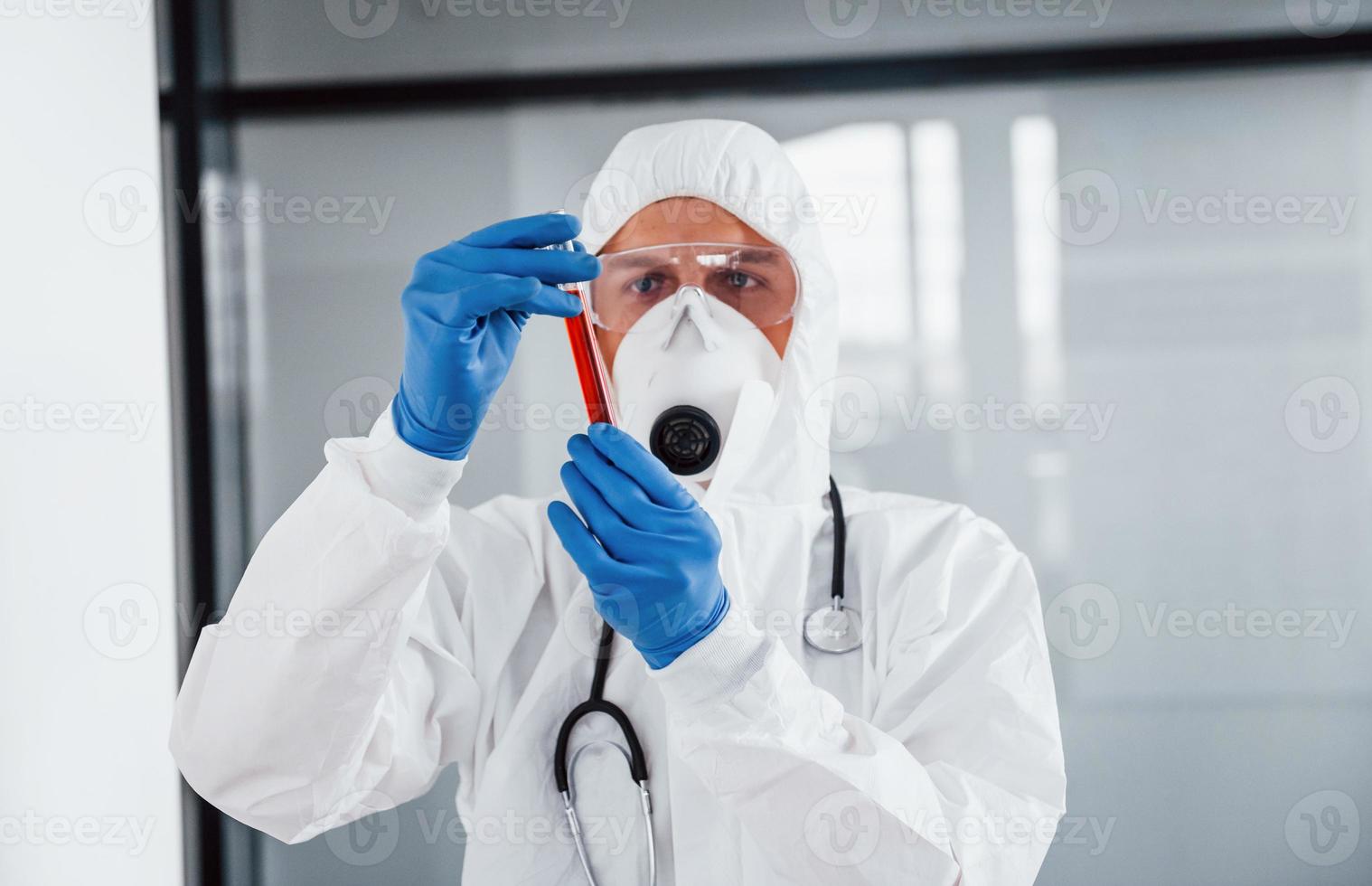 Médecin scientifique de sexe masculin en blouse de laboratoire, lunettes défensives et masque contenant un tube à essai avec du sang photo