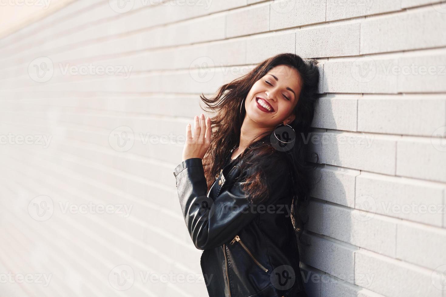 portrait de brune joyeuse aux cheveux bouclés et en vêtements noirs appuyée sur le mur photo