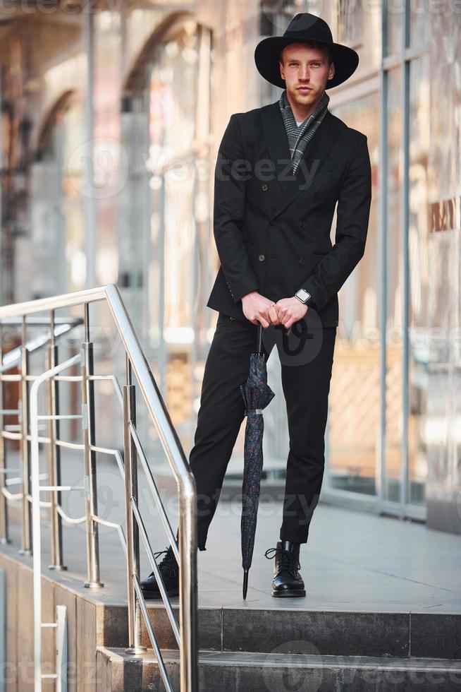 élégant jeune homme au chapeau noir et avec parapluie à l'extérieur de la ville photo