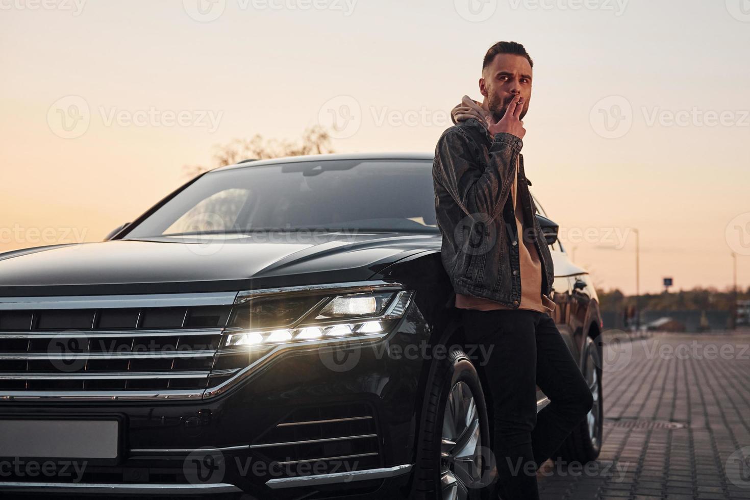 bel homme non rasé dans des vêtements à la mode debout près de sa voiture noire et fumer photo