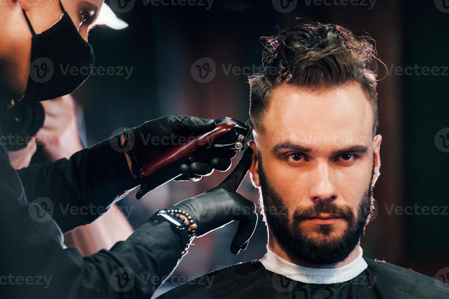 vue de face d'un jeune homme barbu assis et se faisant couper les cheveux dans un salon de coiffure par un gars en masque de protection noir photo