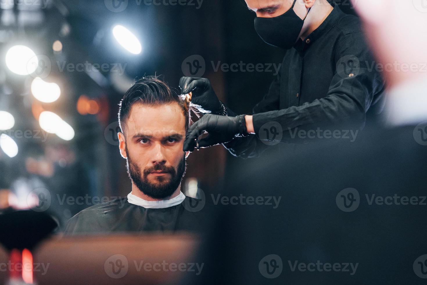 vue de face d'un jeune homme barbu assis et se faisant couper les cheveux dans un salon de coiffure par un gars en masque de protection noir photo