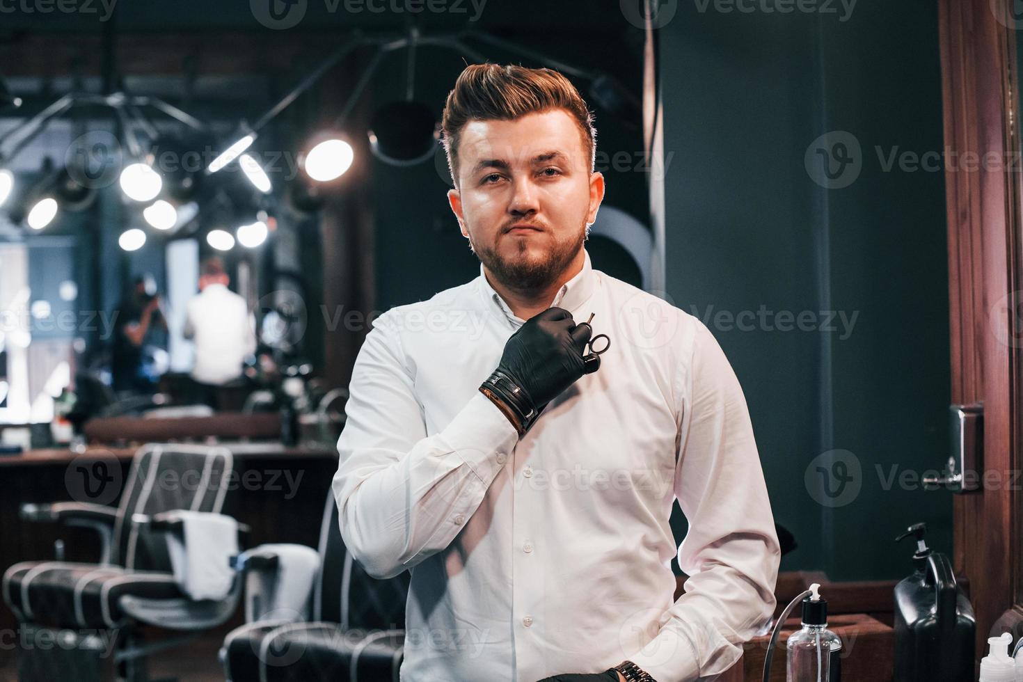 portrait de jeune homme en vêtements blancs et gants noirs qui se tient à l'intérieur dans un salon de coiffure et tenant des ciseaux photo