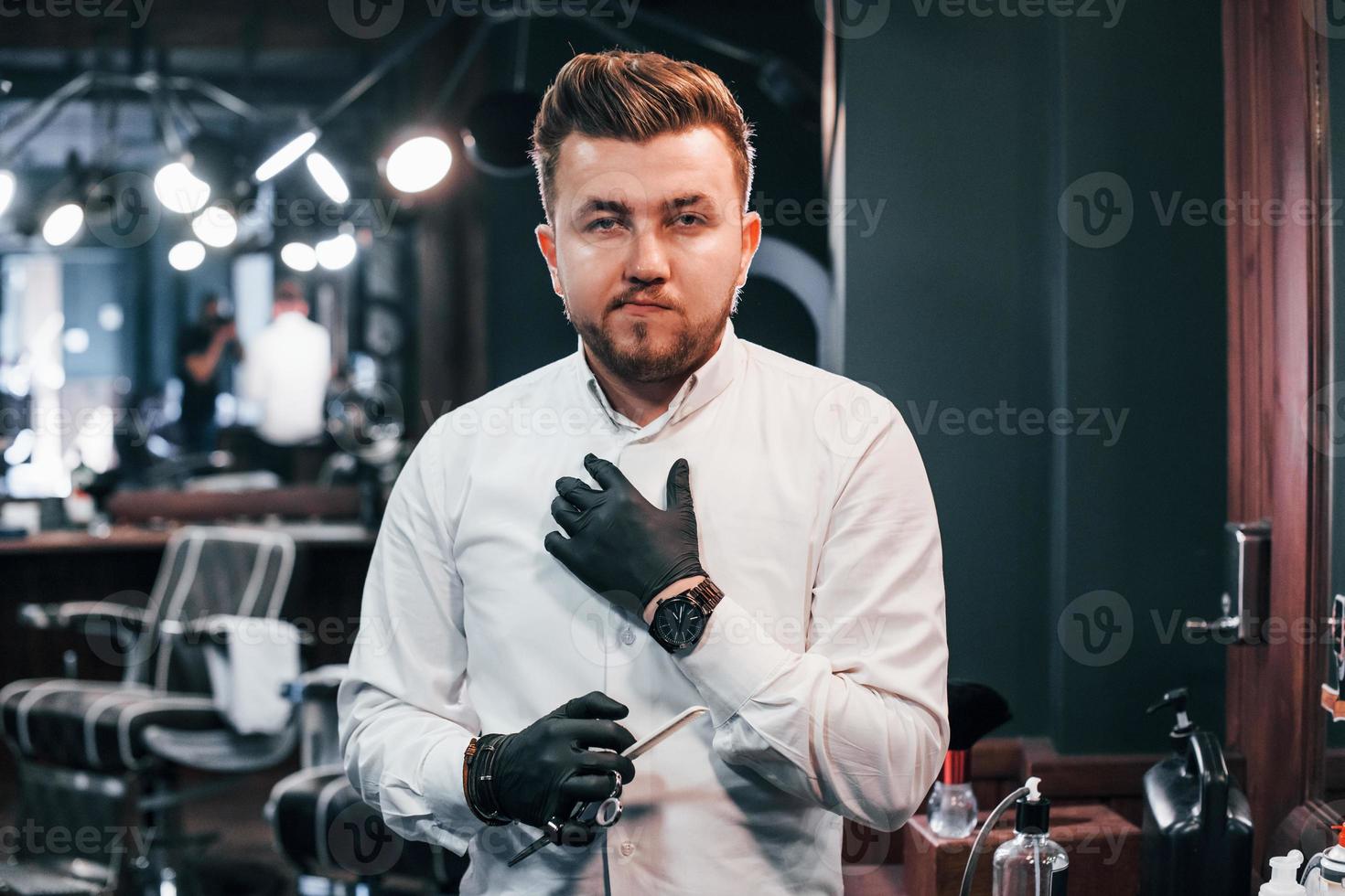 portrait de jeune homme en vêtements blancs et gants noirs qui se tient à l'intérieur dans un salon de coiffure et tenant des ciseaux photo
