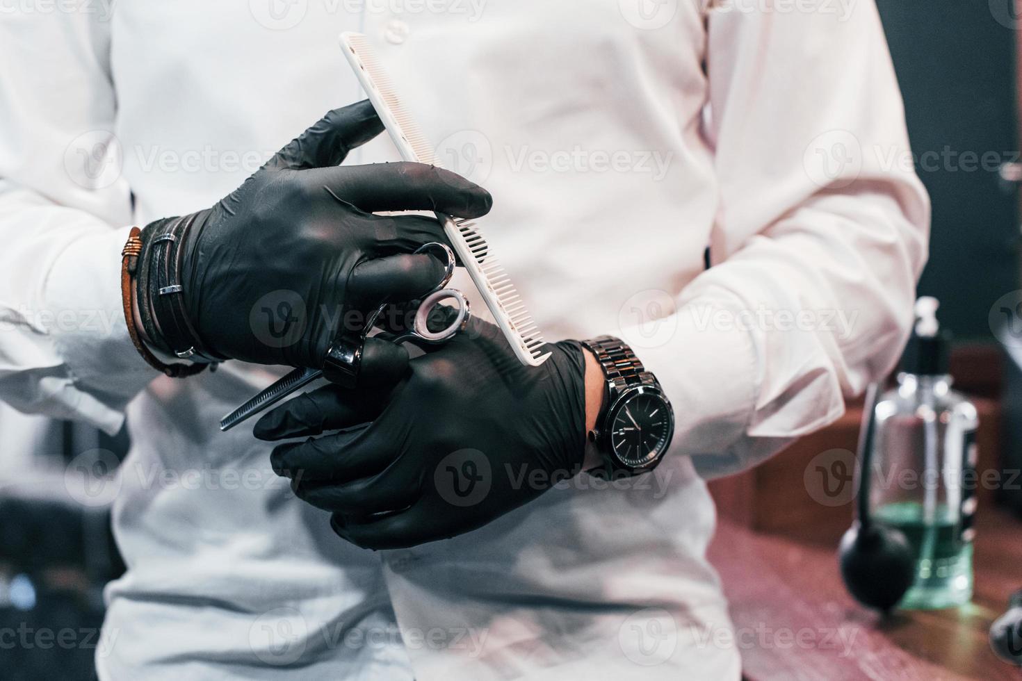 vue rapprochée d'un gars en vêtements blancs et gants noirs qui se tient à l'intérieur dans un salon de coiffure et tenant des ciseaux photo