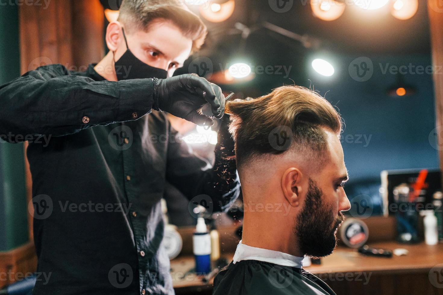 jeune homme barbu assis et se faisant couper les cheveux dans un salon de coiffure par un gars en masque de protection noir photo