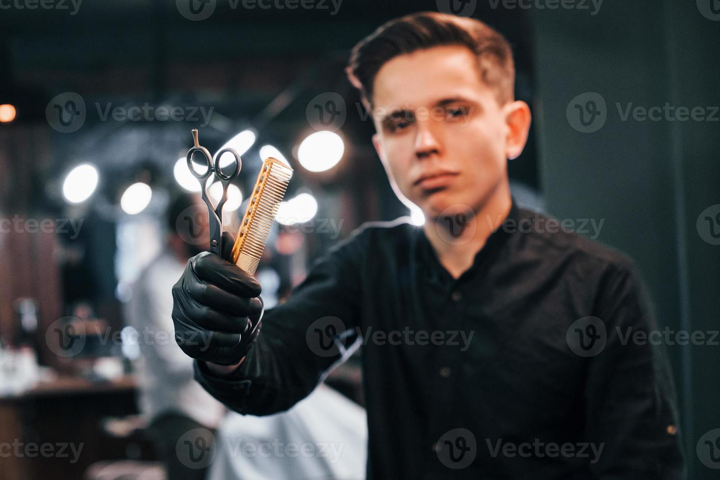 portrait de jeune homme en vêtements noirs qui se tient à l'intérieur dans un salon de coiffure et tenant des ciseaux et un peigne photo