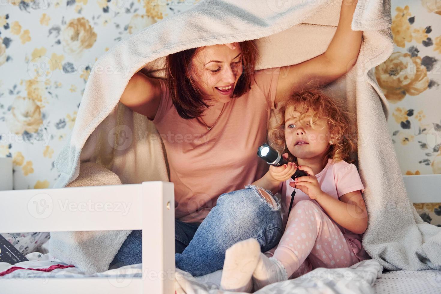 assis sous une couverture avec une lampe de poche. jeune mère avec sa petite fille dans des vêtements décontractés ensemble à l'intérieur à la maison photo