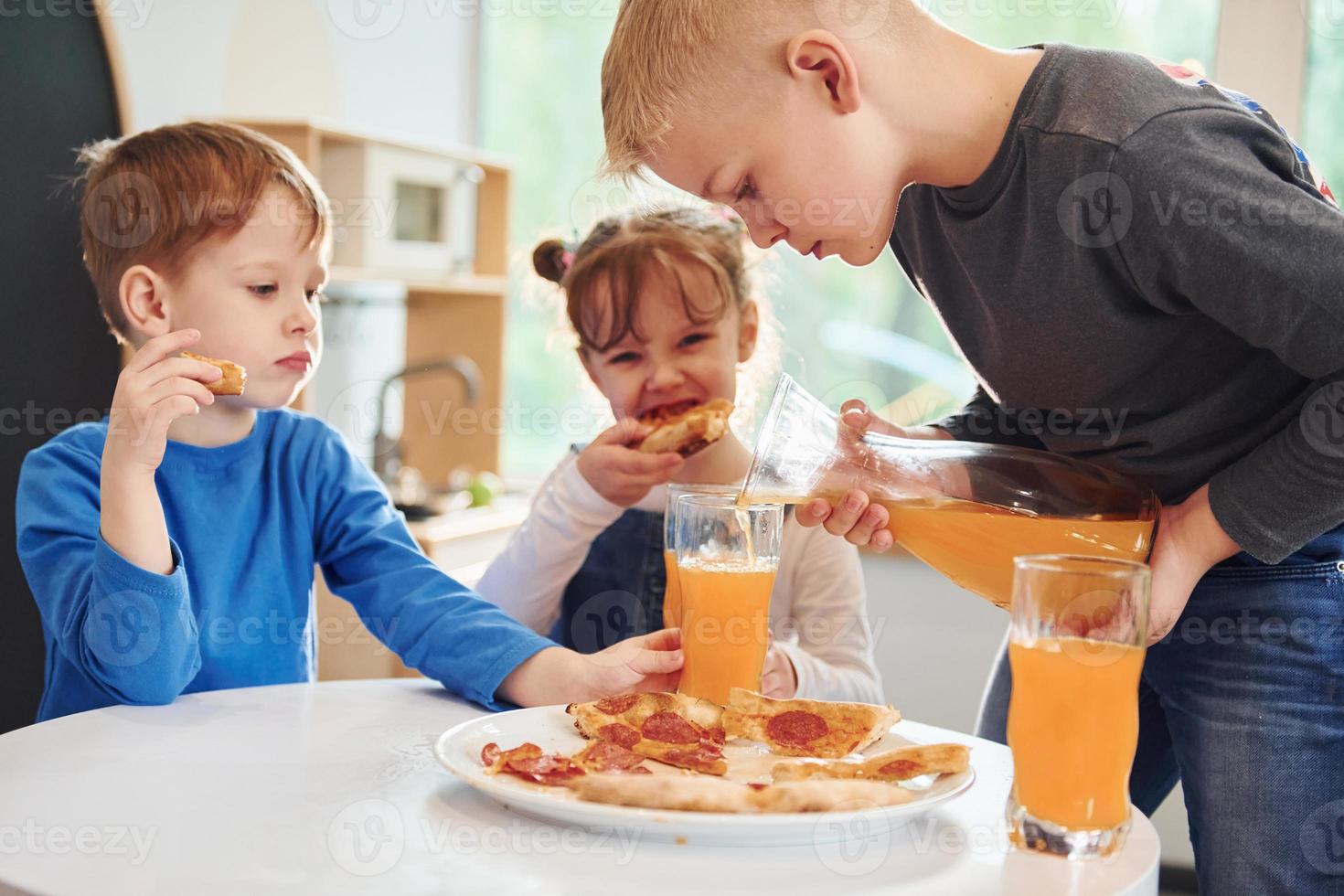 trois enfants assis à l'intérieur près de la table et mangeant ensemble une pizza avec du jus d'orange photo