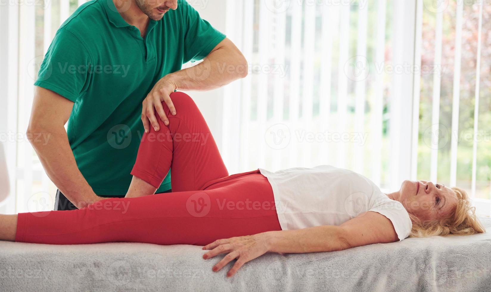 une femme plus âgée a une visite à la clinique, allongée et en rééducation. instructeur masculin l'aidant photo