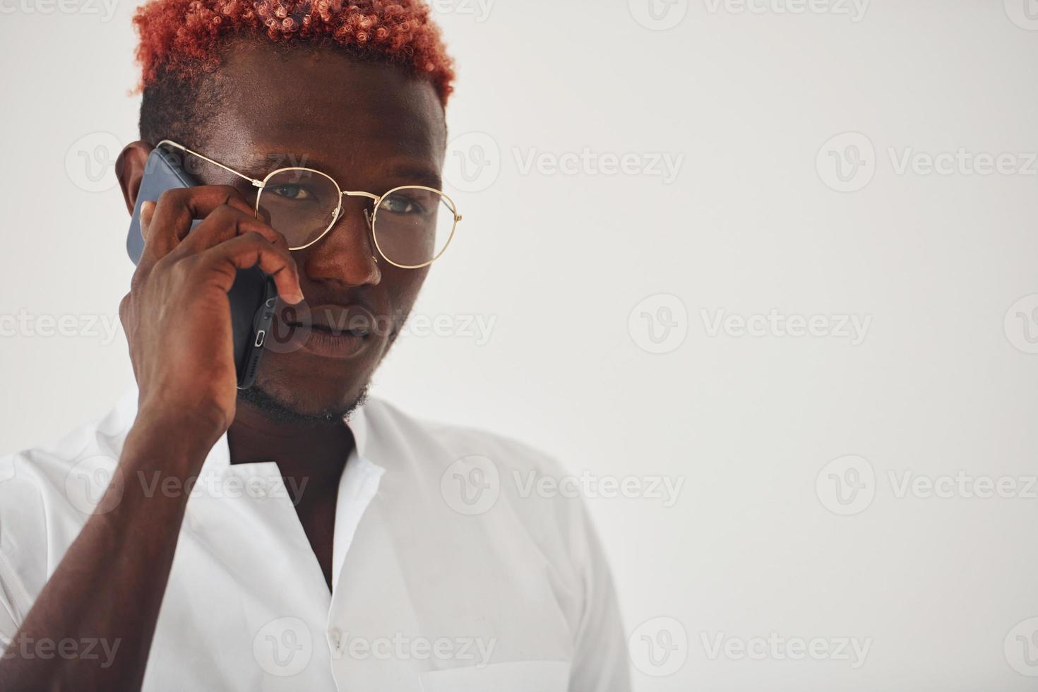 jeune homme afro-américain en vêtements formels blancs parlant au téléphone contre le mur à l'intérieur photo