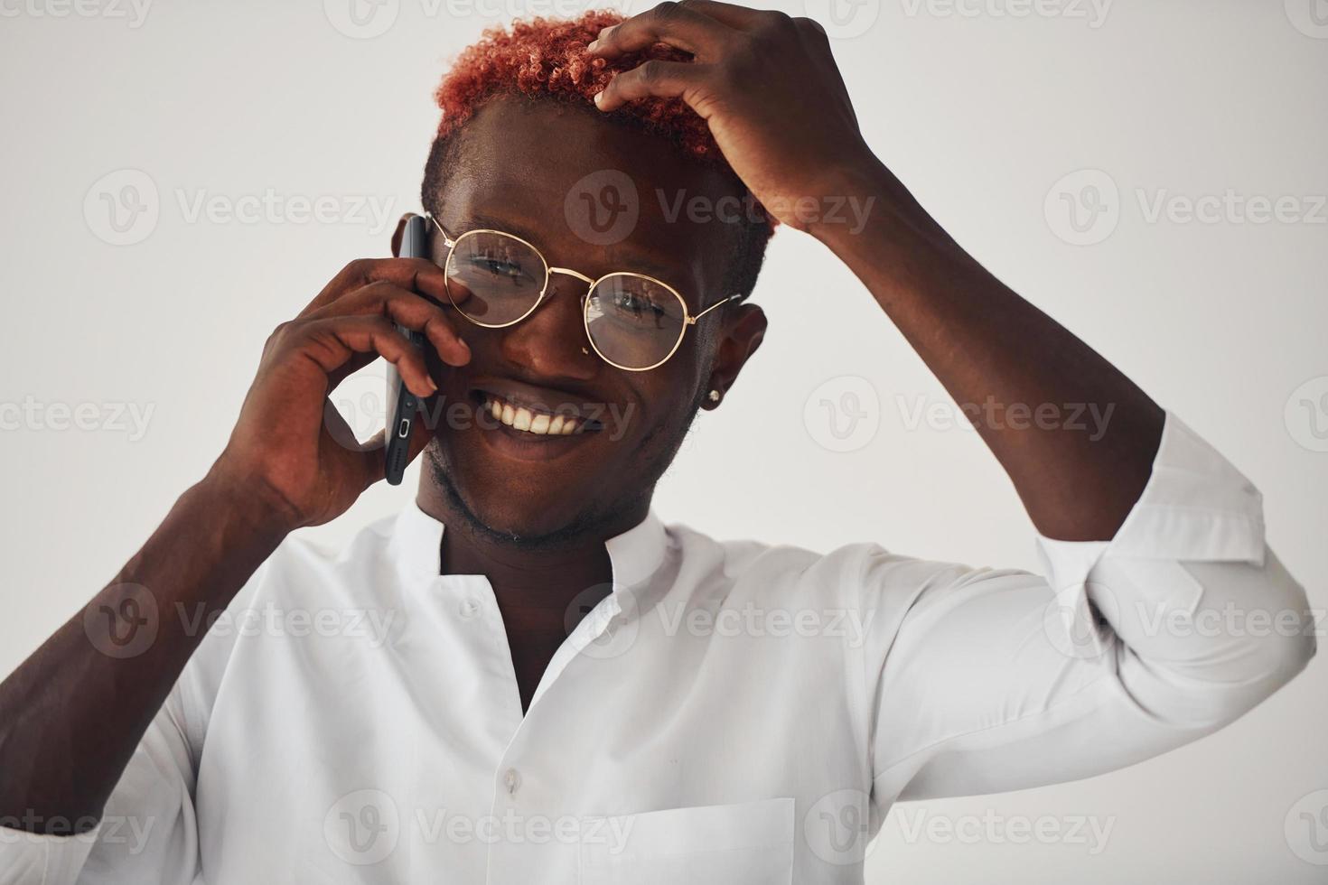 jeune homme afro-américain en vêtements formels blancs parlant au téléphone contre le mur à l'intérieur photo