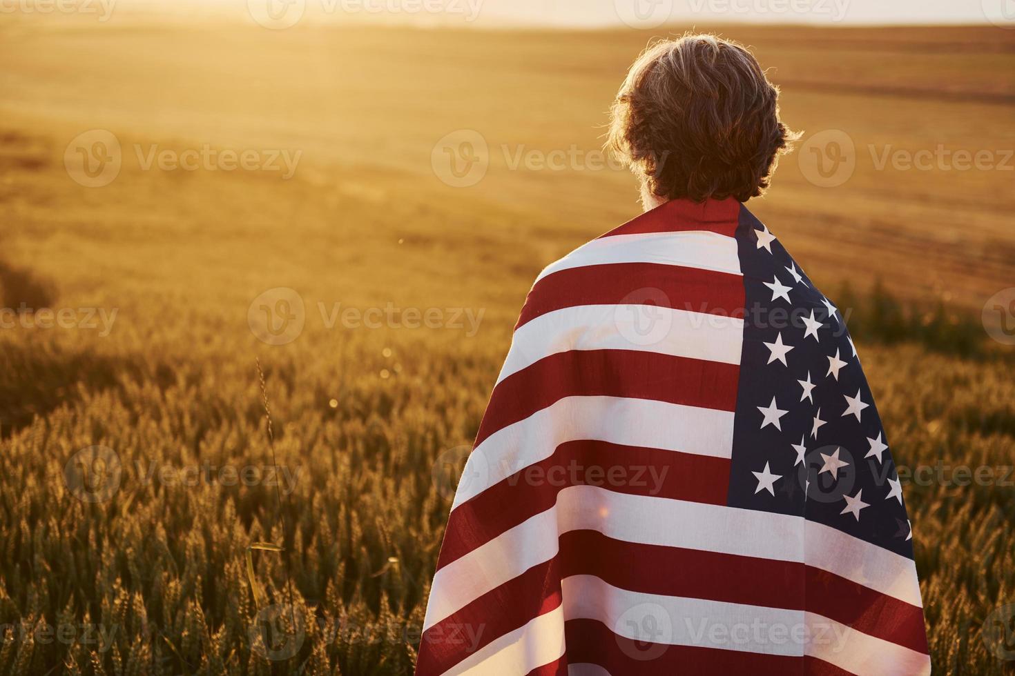 vue de derrière. tenant le drapeau américain dans les mains. homme élégant senior patriotique aux cheveux gris et à la barbe sur le terrain agricole photo