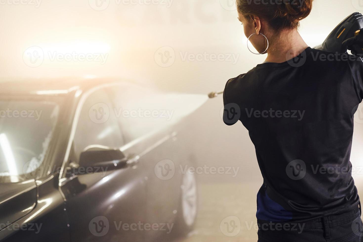 utiliser des équipements avec de l'eau à haute pression. une automobile noire moderne est nettoyée par une femme à l'intérieur d'une station de lavage de voiture photo