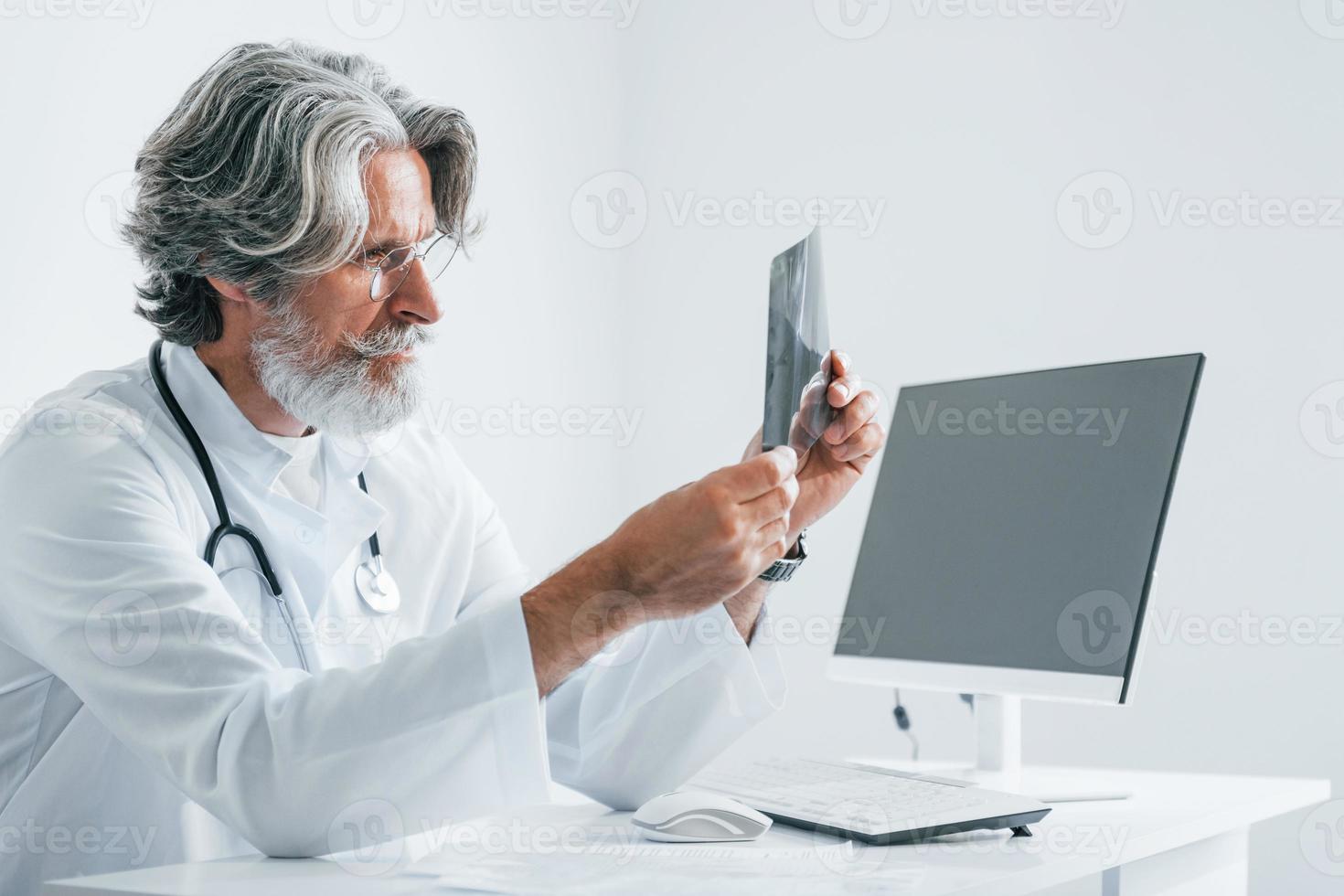 regarde la radiographie. médecin senior aux cheveux gris et à la barbe en blouse blanche est à l'intérieur de la clinique photo