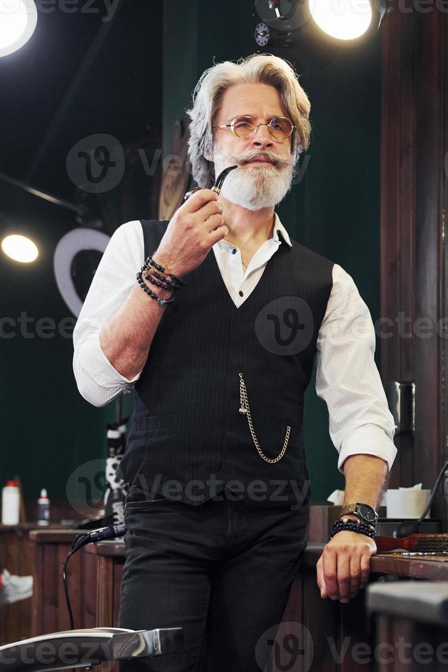 debout et fumer dans un salon de coiffure. homme âgé moderne et élégant aux cheveux gris et à la barbe est à l'intérieur photo