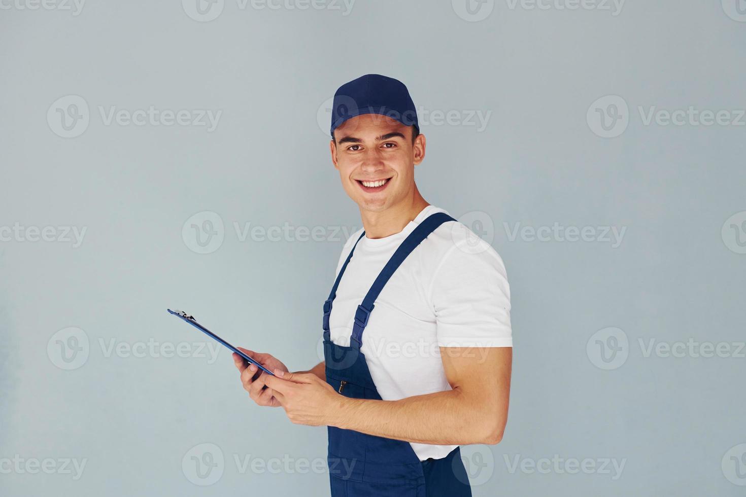 en casquette et avec bloc-notes. travailleur masculin en uniforme bleu debout à l'intérieur du studio sur fond blanc photo