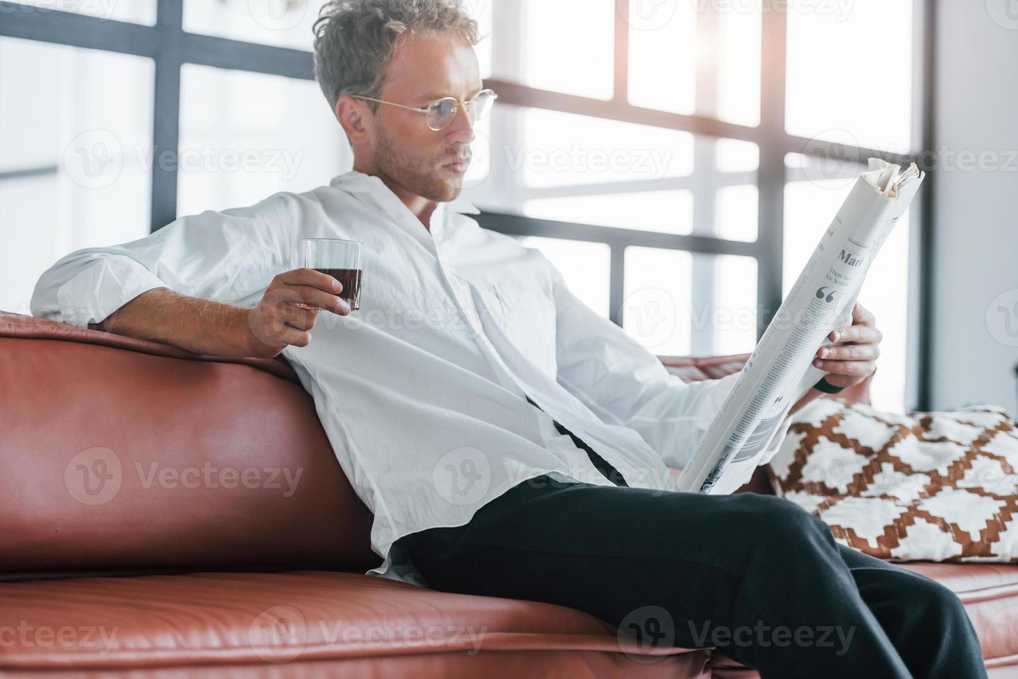 lit le journal. jeune homme caucasien en chemise blanche élégante à l'intérieur à la maison photo