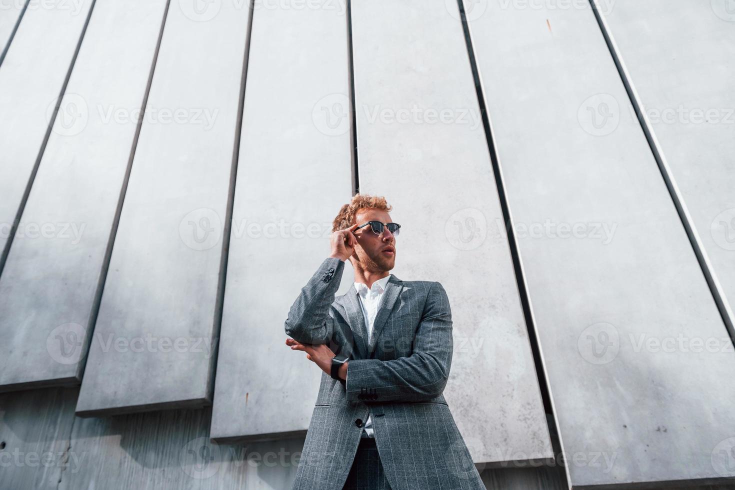 debout contre le mur du bâtiment. jeune homme d'affaires en tenue de soirée grise est à l'extérieur de la ville photo