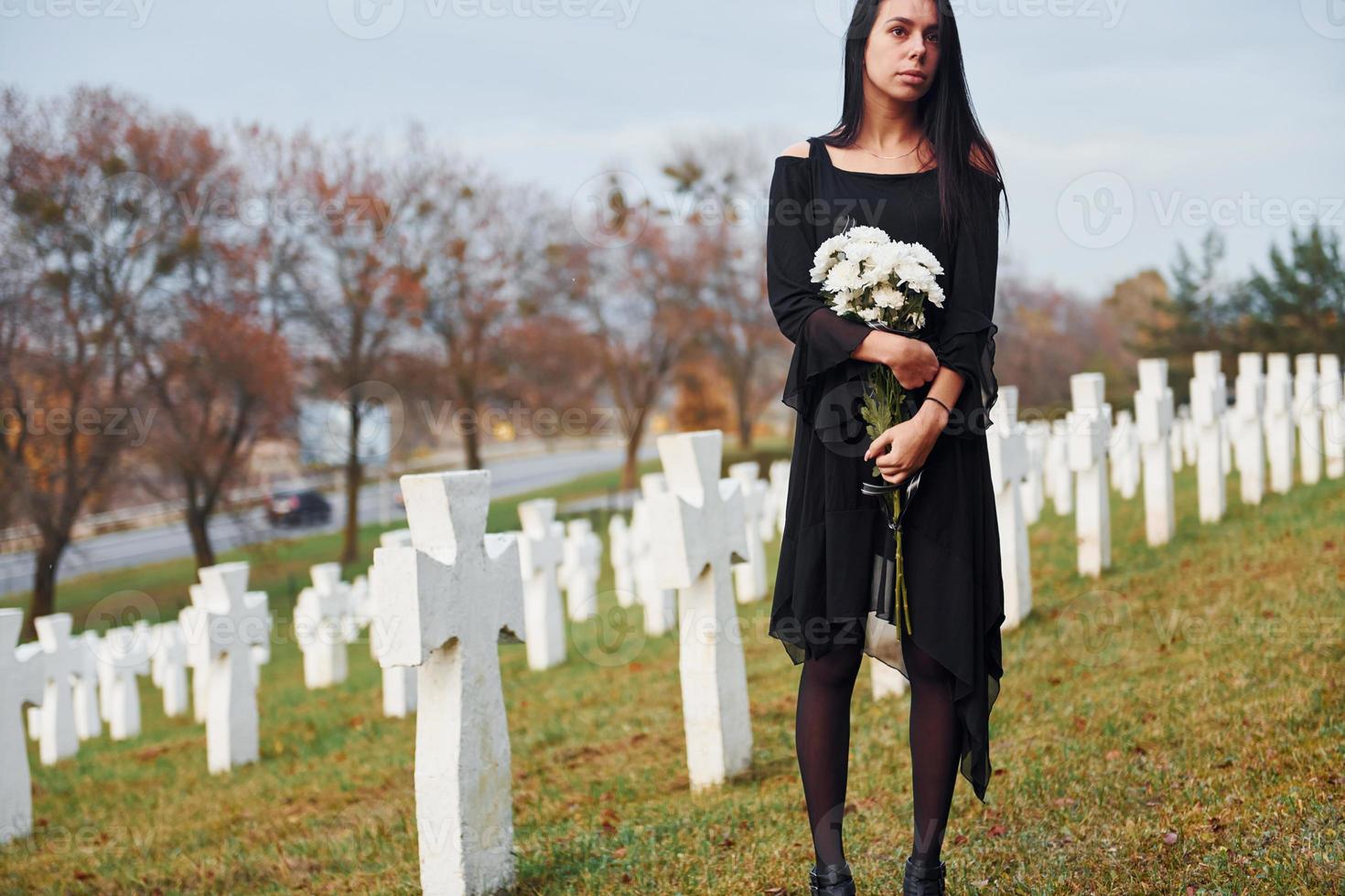 avec des fleurs dans les mains. jeune femme en vêtements noirs visitant le cimetière avec de nombreuses croix blanches. conception des funérailles et de la mort photo