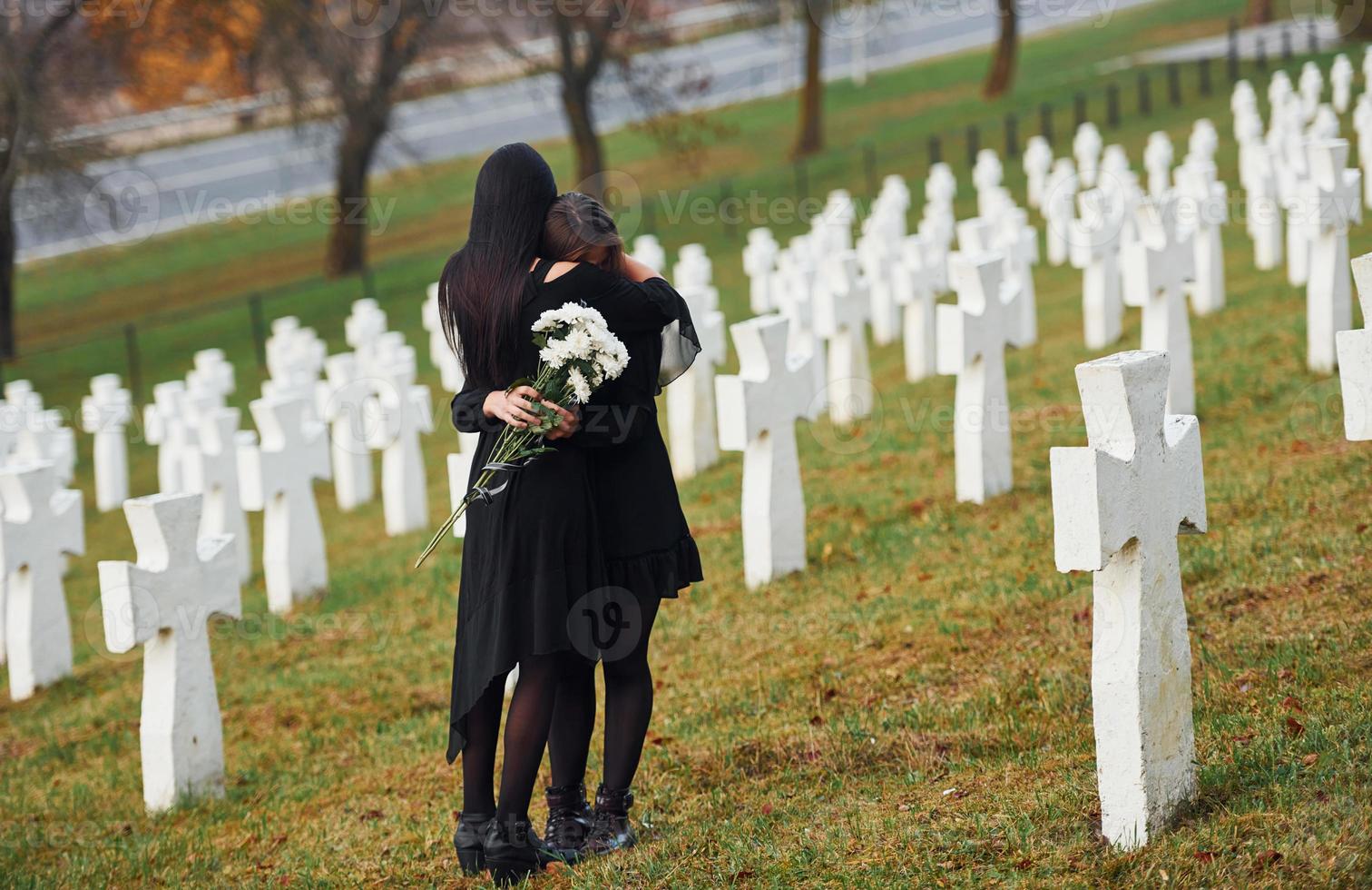 s'embrassent et pleurent. deux jeunes femmes en vêtements noirs visitant un cimetière avec de nombreuses croix blanches. conception des funérailles et de la mort photo