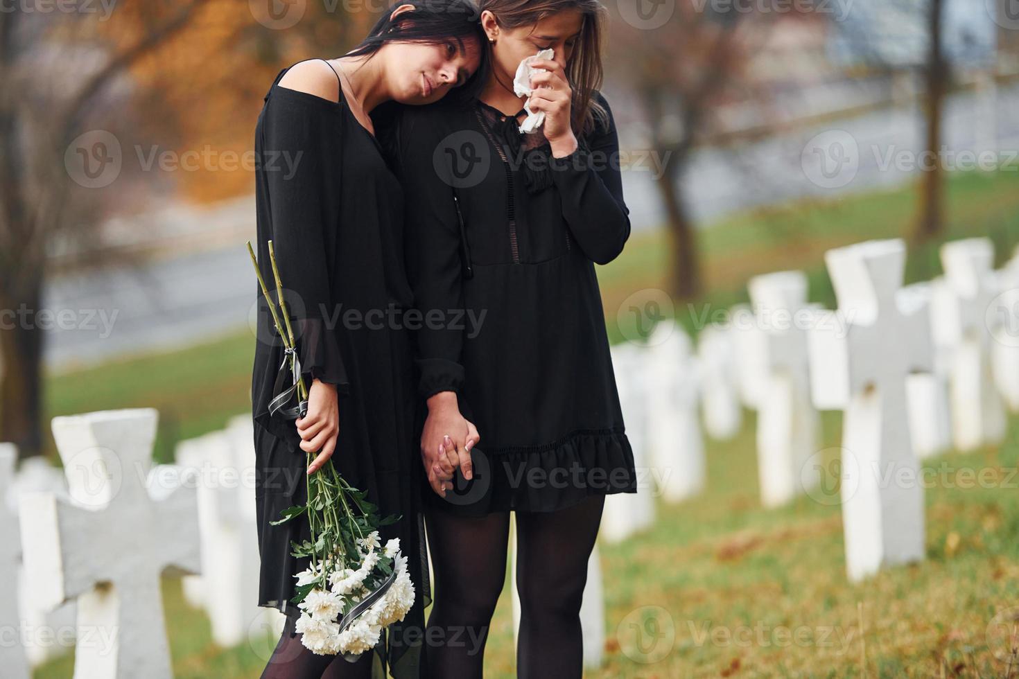 détient des fleurs. deux jeunes femmes en vêtements noirs visitant un cimetière avec de nombreuses croix blanches. conception des funérailles et de la mort photo
