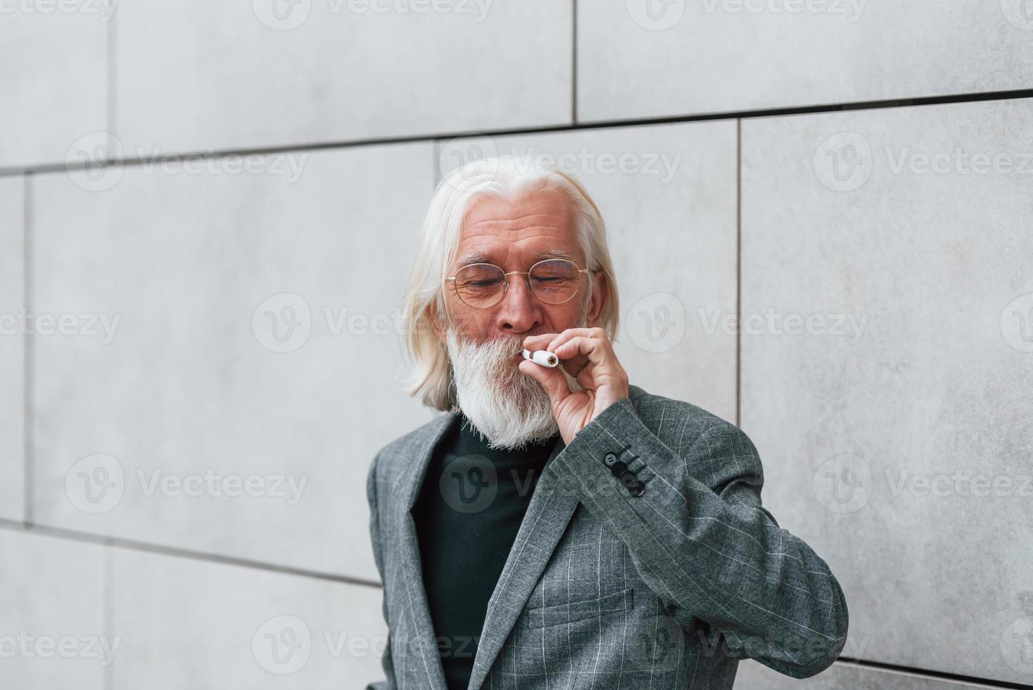 homme d'affaires senior en vêtements formels, aux cheveux gris et à la barbe est à l'extérieur fume une cigarette électronique photo