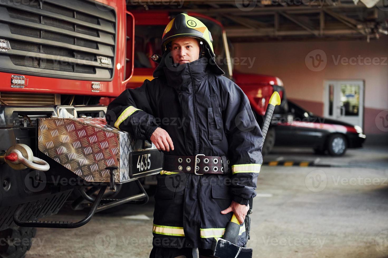 tient la hache dans les mains. pompier masculin en uniforme de protection debout près du camion photo