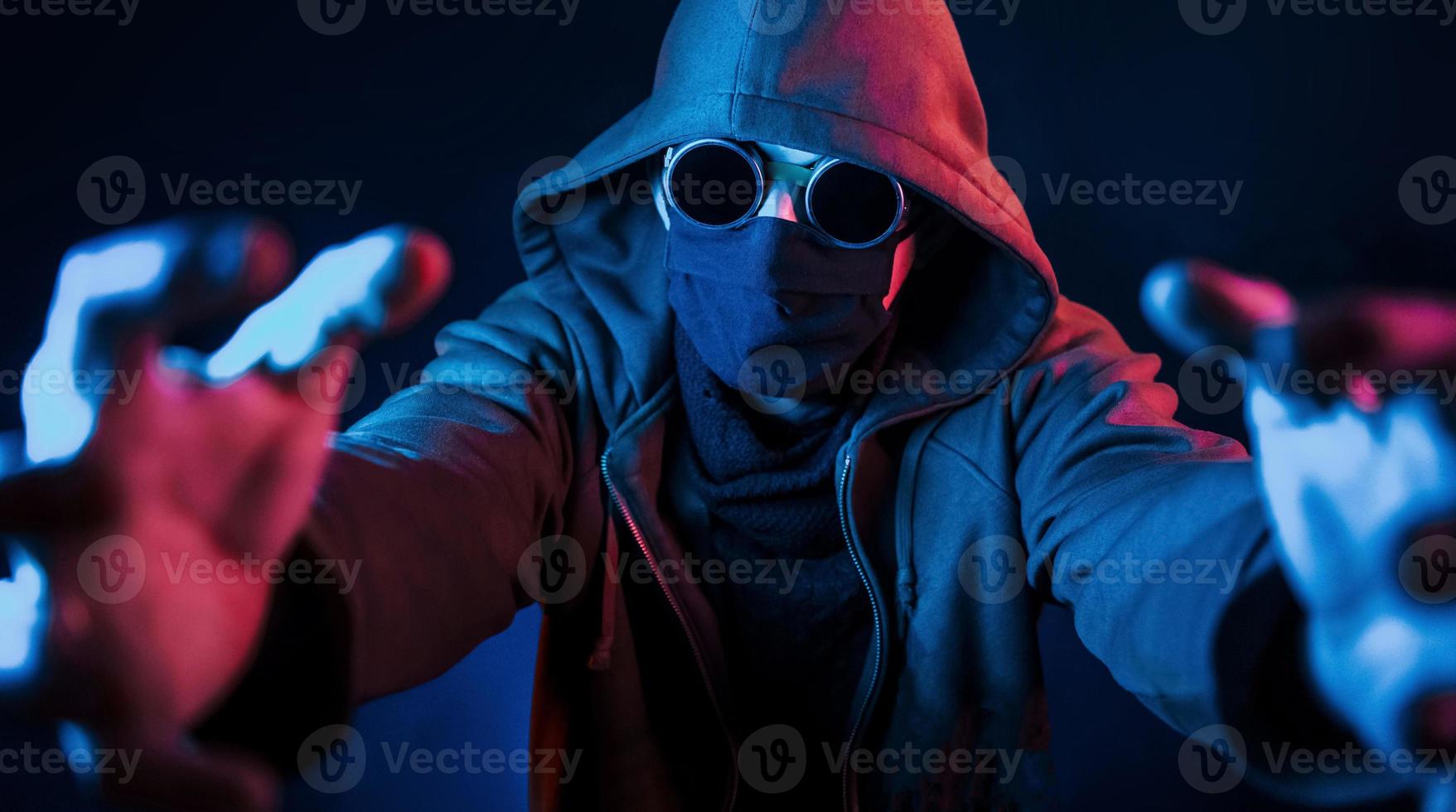 mec effrayant en lunettes, masque et cagoule. éclairage au néon. jeune homme européen est dans le studio sombre photo