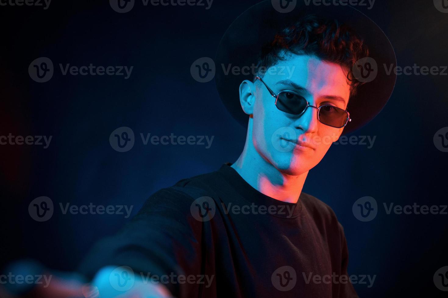 en chapeau et lunettes. éclairage au néon. jeune homme européen est dans le studio sombre photo