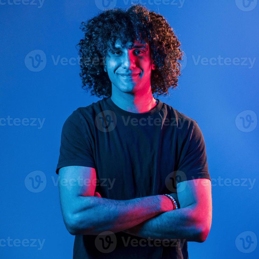 posant pour un appareil photo. jeune bel homme aux cheveux bouclés est à l'intérieur dans le studio avec éclairage au néon photo