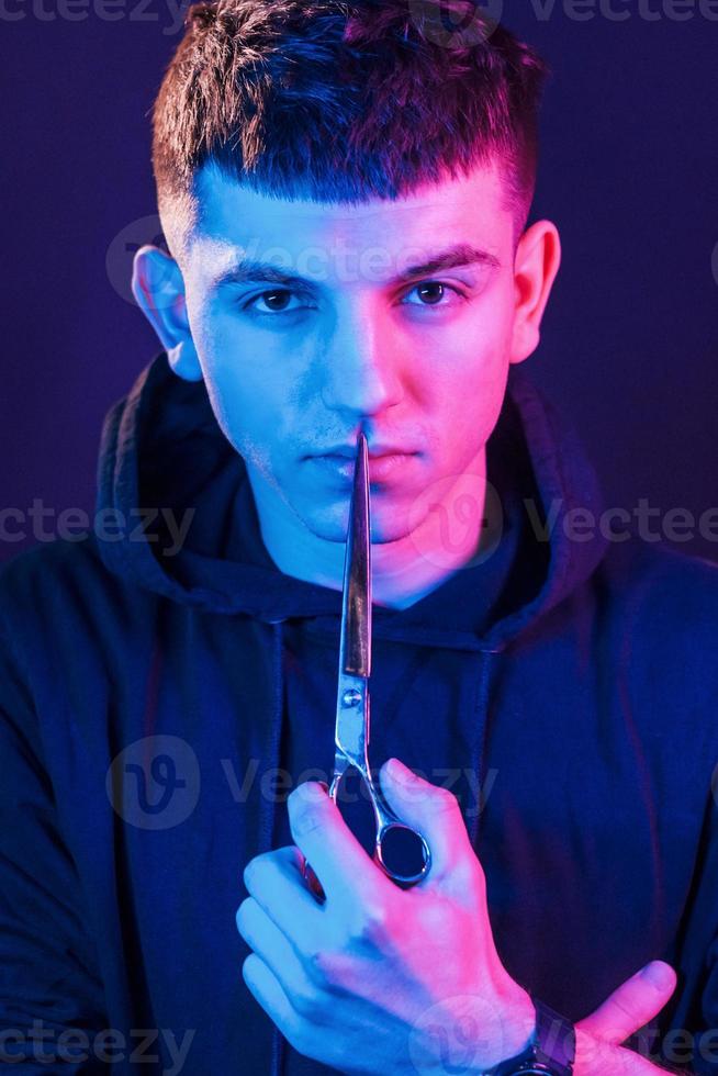 tient des ciseaux. jeune coiffeur avec équipement de travail debout dans le studio avec éclairage au néon photo