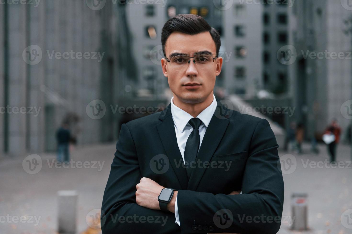 debout les bras croisés. homme d'affaires en costume noir et cravate est à l'extérieur de la ville photo