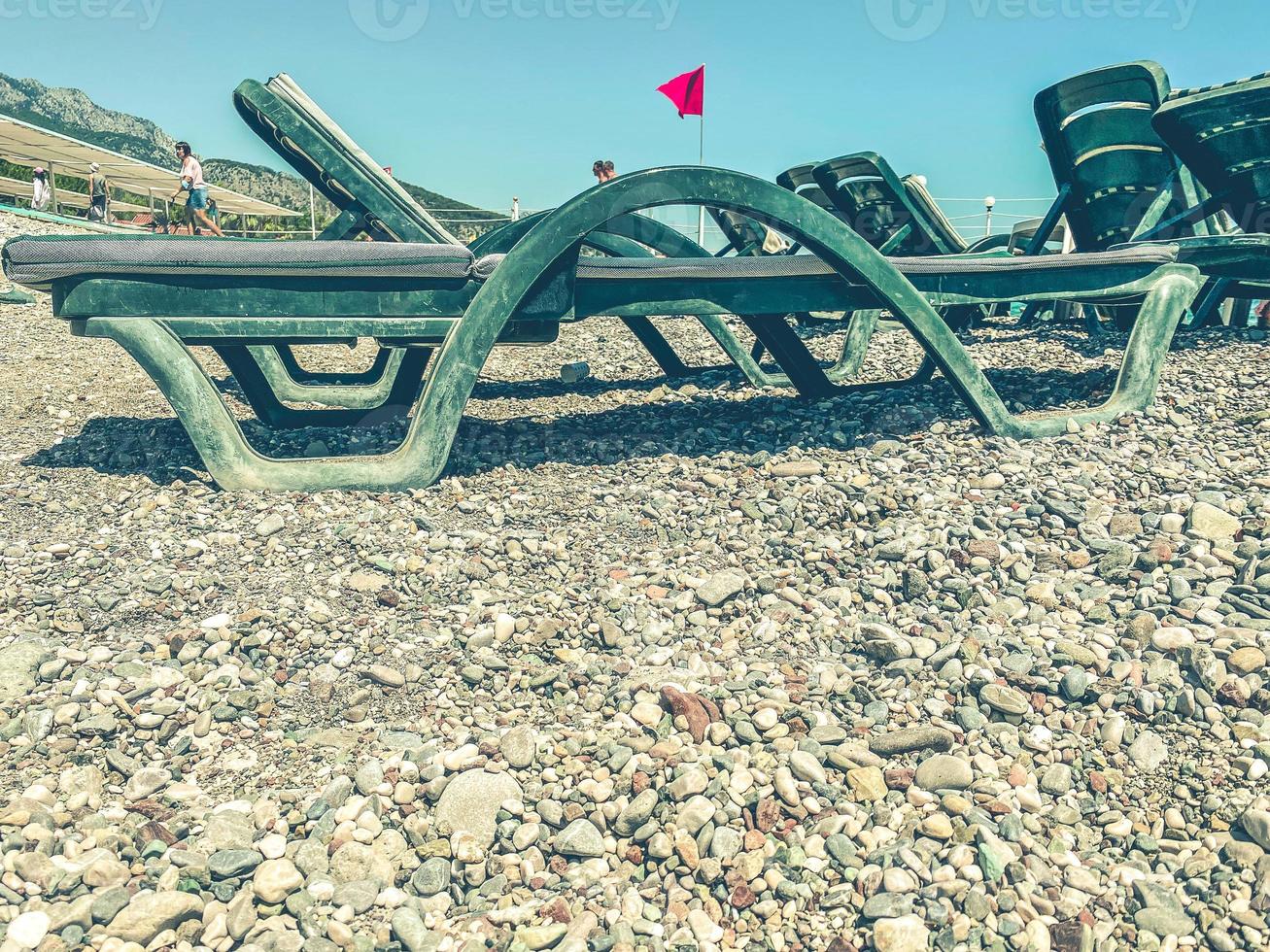 vacances au bord de la mer sur la plage. sur le sol caillouteux, il y a des chaises longues pour les touristes qui prennent le soleil. il y a des matelas et du tissu vert sur les transats. sur le fond du drapeau turc photo