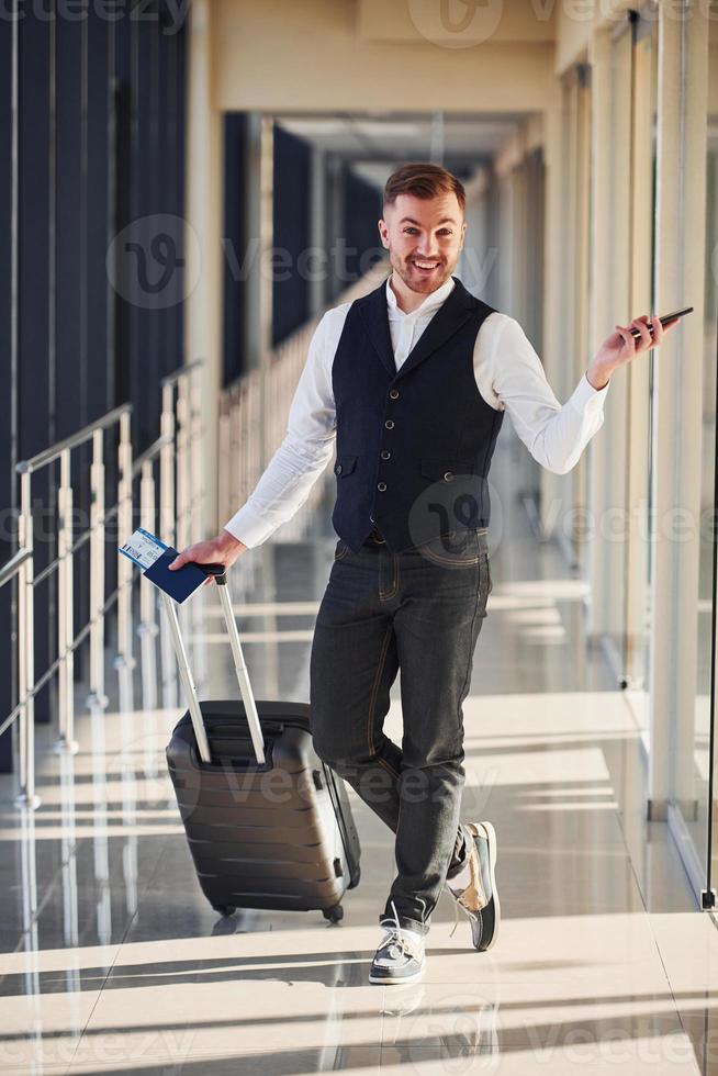 un passager masculin vêtu d'élégants vêtements de cérémonie se trouve dans le hall de l'aéroport avec des bagages, des billets et un téléphone photo