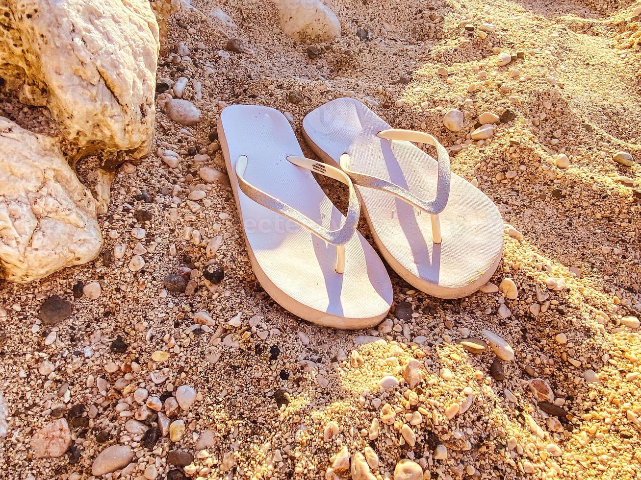 des pantoufles en caoutchouc blanc se tiennent sur le sable. chaussures de mer. ardoises pour se promener le long de la côte maritime. pantoufles pour femmes qui se portent sur un doigt photo