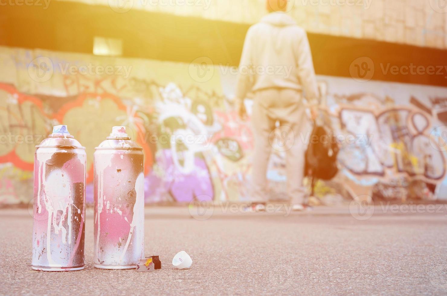 plusieurs bombes aérosols usagées avec de la peinture rose et blanche se trouvent sur l'asphalte contre le gars debout devant un mur peint dans des dessins de graffitis colorés photo