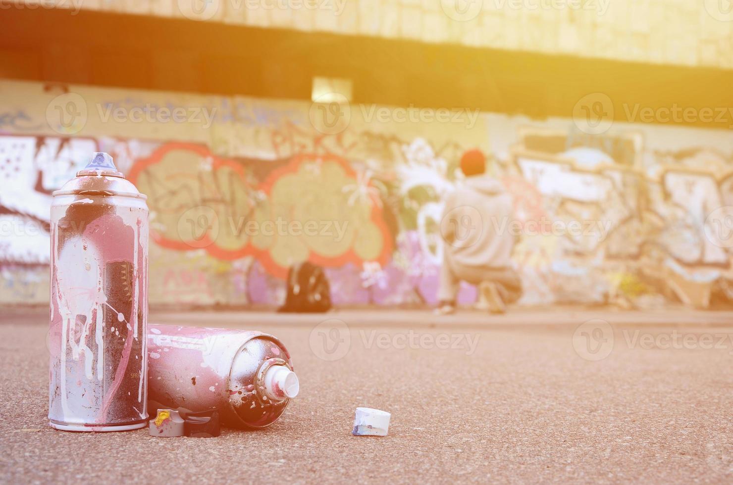 plusieurs bombes aérosols usagées avec de la peinture rose et blanche se trouvent sur l'asphalte contre le gars debout devant un mur peint dans des dessins de graffitis colorés photo