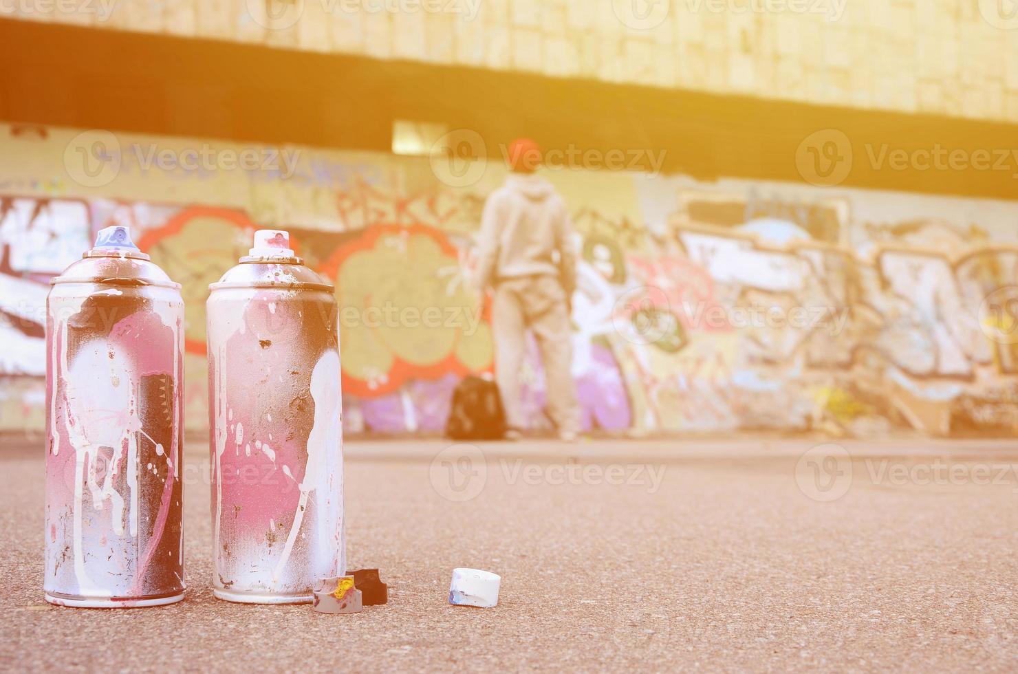 plusieurs bombes aérosols usagées avec de la peinture rose et blanche se trouvent sur l'asphalte contre le gars debout devant un mur peint dans des dessins de graffitis colorés photo