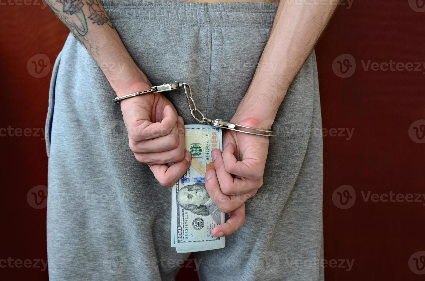 un gars arrêté en pantalon gris avec des mains menottées détient une énorme quantité de billets d'un dollar. vue arrière photo