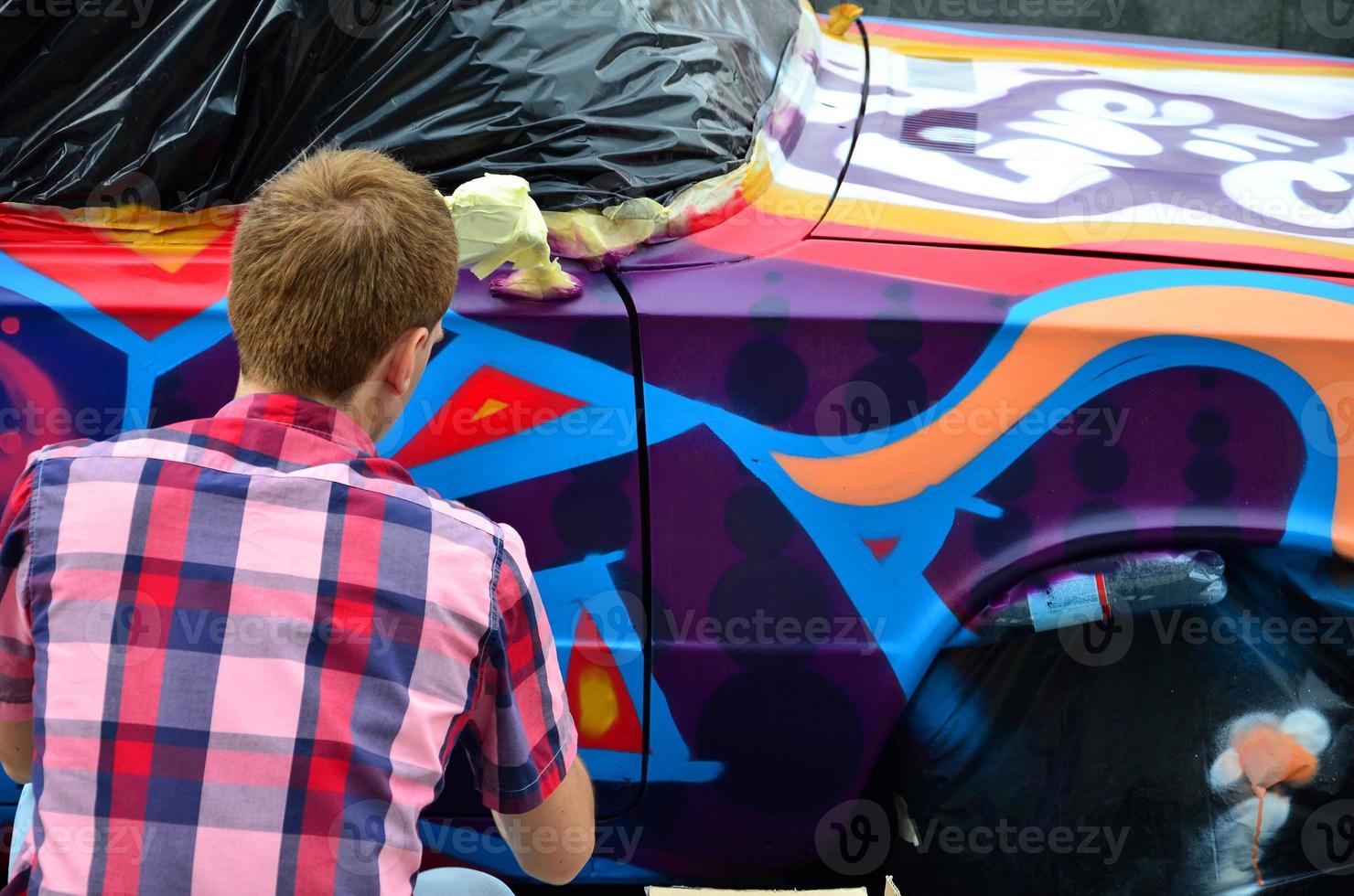 un jeune graffeur aux cheveux roux peint un nouveau graffiti coloré sur la voiture. photo du processus de dessin d'un graffiti sur un gros plan de voiture. le concept d'art de rue et de vandalisme illégal