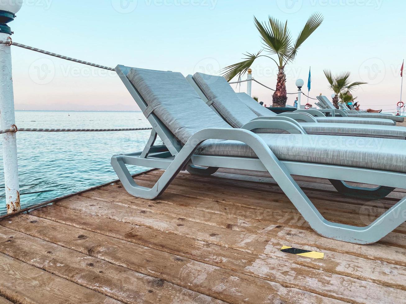 pays chaud et tropical avec mer et montagnes. ponton en bois pour plonger dans l'eau. au sol il y a un transat en tissu blanc à côté d'un palmier pour se protéger du soleil photo