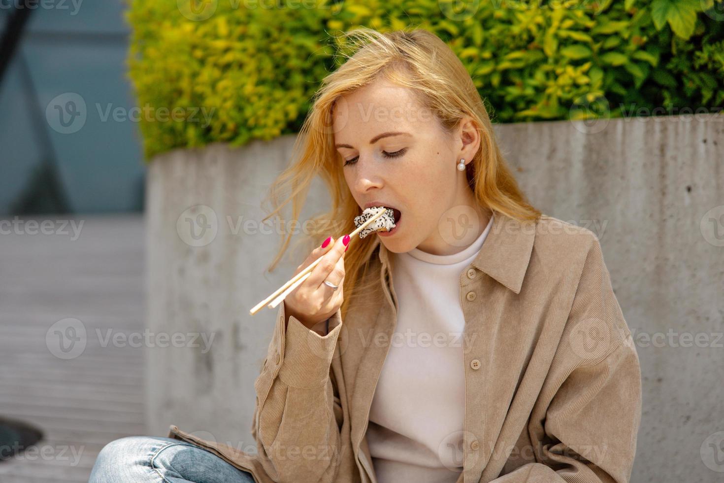 jeune belle femme blonde mangeant des sushis à l'extérieur, sur la terrasse en bois, par un bâtiment moderne de la ville. nourriture savoureuse à emporter. la fille a une pause déjeuner, passe du temps à l'extérieur et mange de la nourriture asiatique. la vie en ville photo