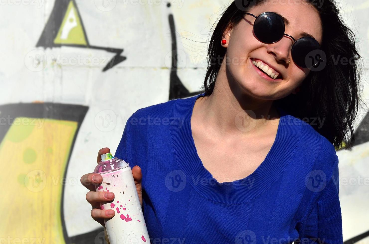 portrait d'une jeune fille émotionnelle aux cheveux noirs et aux piercings. photo d'une fille avec des pots de peinture aérosol dans les mains sur un fond de mur de graffiti. le concept de street art et l'utilisation de peintures aérosols